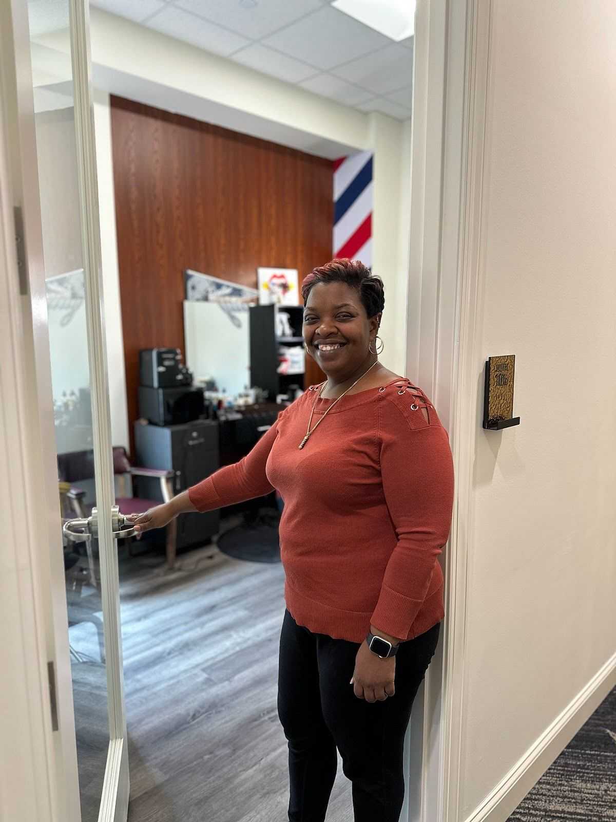 Person smiling, opening door to salon with barber's pole in background.